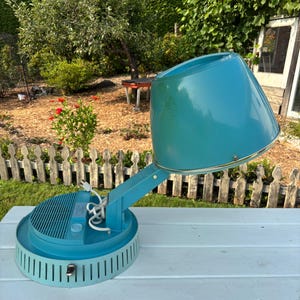 May include: Vintage teal hair dryer with a conical hood and a round base. The base has a series of vertical slots around the perimeter. The dryer is on a white surface, with a garden in the background.