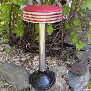 May include: A retro-style bar stool with a red padded seat and chrome accents. The stool has a chrome pole and a black, circular base. The seat is trimmed with chrome and red detailing, evoking a classic diner aesthetic.
