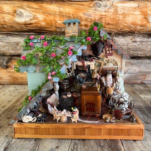 May include: A detailed miniature house diorama with a wooden base and roof. The roof is decorated with pink roses and green vines. The house is filled with miniature furniture and objects, creating a whimsical scene. The diorama is set on a wooden surface.
