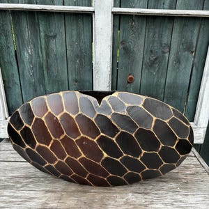 Könnte beinhalten: Eine große, ovale Holzschale mit einem geometrischen Muster in Dunkelbraun und Hellbraun. Die Schale ist mit einem Wabenmuster verziert und hat einen leicht erhöhten Rand.