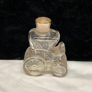 Puede incluir: Una botella de vidrio transparente con forma de coche antiguo con una tapa dorada. La botella tiene un diseño detallado con ruedas y un carruaje.