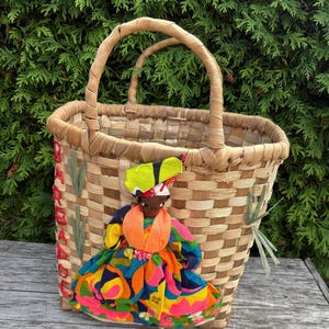 May include: Woven basket with a doll attached. The basket has a rectangular shape with a woven pattern and two handles. The doll is wearing a colorful dress with orange, blue, and pink accents. The basket is decorated with red and green accents.