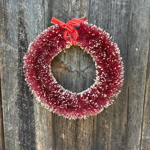May include: A red and white Christmas wreath with a red ribbon bow. The wreath is made of a fluffy material and is hanging on a wooden door.