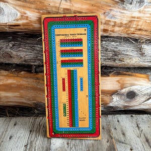 Könnte beinhalten: Cribbage-Brett aus Holz mit durchgehendem Streckendesign. Das Brett hat rote, grüne und blaue Bahnen mit Löchern für die Stifte. Der Text auf dem Brett enthält "Continuous Track Cribbage" und Etiketten für "Legs", "Skunks" und "Total Points."