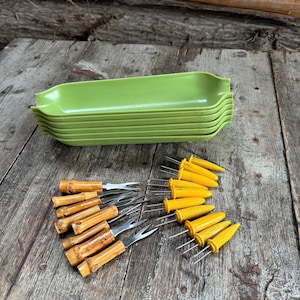 May include: A collection of green, corn-shaped serving dishes stacked on a wooden surface. Bamboo-handled corn holders and yellow corn-shaped holders are also present, ideal for a summer barbecue or picnic.