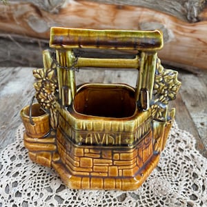 May include: A brown and green ceramic well planter with a metal chain and bucket. The well has a brick design and the word "AWISH" written on the front.