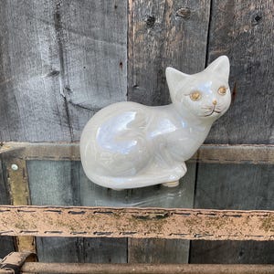 May include: A white ceramic cat figurine with gold eyes. The cat is sitting with its head turned to the side and its tail curled around its body.