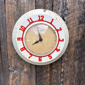 Op de afbeelding: Een vintage, ronde klok met een witte wijzerplaat en rode cijfers. De wijzerplaat heeft een beige achtergrond met zwarte wijzers en de tekst "8 DAY". De klok is gemonteerd op een verweerde houten muur.