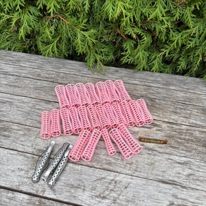 May include: A collection of pink hair rollers and silver hair clips arranged on a wooden surface. The pink rollers are cylindrical with a lattice design. The background features green foliage.