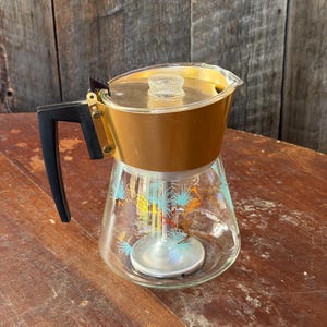 May include: A glass percolator coffee pot featuring a gold-toned metal upper band, a black plastic handle, and a clear glass base decorated with a pattern of teal and gold pine needle motifs. The lid includes a clear glass knob.