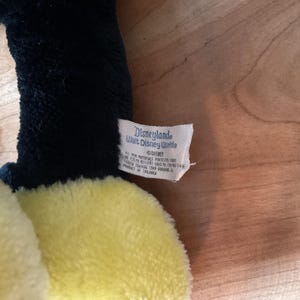May include: A soft plush toy with a yellow body and black detailing. The tag reads "Disneyland Walt Disney World" and features the Disney logo. The toy is made of a soft, textured fabric.