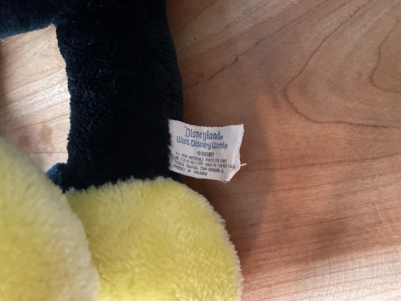 May include: A soft plush toy with a yellow body and black detailing. The tag reads "Disneyland Walt Disney World" and features the Disney logo. The toy is made of a soft, textured fabric.