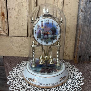 Puede incluir: Un reloj decorativo Thomas Kinkade con una cúpula de cristal. La esfera del reloj presenta una pintura paisajística. La base blanca tiene detalles dorados. El reloj tiene detalles metálicos dorados y un péndulo decorativo.