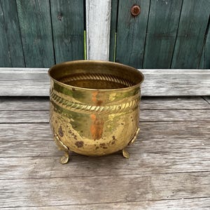 May include: A gold-coloured metal planter with a decorative rope design around the rim. The planter has three legs and is sitting on a wooden surface.