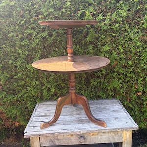 May include: A two-tiered wooden stand featuring a round top shelf and a larger round bottom shelf. The stand is supported by a central turned wooden pillar and rests on a tripod base with three curved legs, all finished in a medium brown wood tone.
