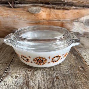 May include: A vintage white casserole dish with a clear glass lid. The dish has a floral pattern in orange, brown, and black. It features two handles and is likely made of heat-resistant glass. A classic kitchenware item.