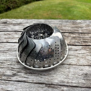 Puede incluir: Un vaporizador de verduras de acero inoxidable, abierto y listo para usar. El vaporizador tiene una base perforada y lados curvos en forma de pétalo. Se encuentra sobre una superficie de madera desgastada, con un fondo verde borroso.