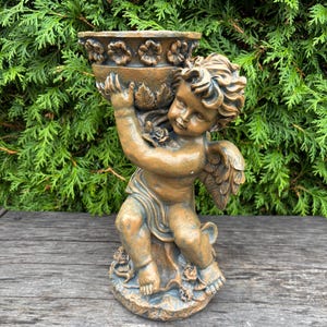 Könnte beinhalten: Eine bronzefarbene Cherub-Statue, die eine dekorative Schale hält. Der Cherub hat detaillierte Flügel und sitzt auf einem mit Rosen geschmückten Sockel. Die Statue steht vor einem Hintergrund aus grünem Laub, was auf eine Gartenumgebung im Freien hindeutet.
