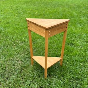 May include: A light brown wooden corner table with a triangular top and a matching triangular lower shelf. The table features three straight legs and visible joinery details on the upper frame, constructed from smooth, finished wood.