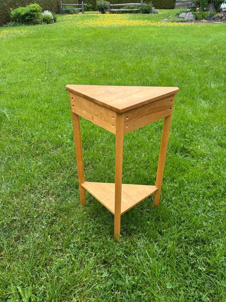 May include: A light brown wooden corner table with a triangular top and a matching triangular lower shelf. The table features three straight legs and visible joinery details on the upper frame, constructed from smooth, finished wood.
