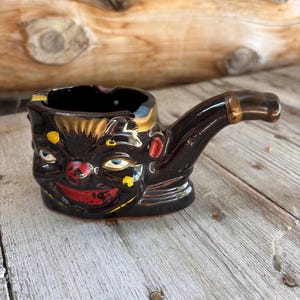 Puede incluir: Un cenicero de cerámica vintage con forma de cabeza de payaso. El cenicero es principalmente negro con detalles pintados que incluyen una nariz y boca rojas, ojos azules y detalles amarillos. El mango en forma de pipa se extiende desde el lateral.