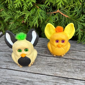Könnte beinhalten: Zwei Vintage-Furby-Spielzeuge. Eines ist cremefarben mit schwarz-weißen Ohren, einem schwarzen Schnabel und grünen Augen. Das andere ist gelb mit großen gelben Ohren, rosa Augen und einem orangefarbenen Schnabel. Beide haben einen kleinen Büschel farbigen Fells auf dem Kopf.