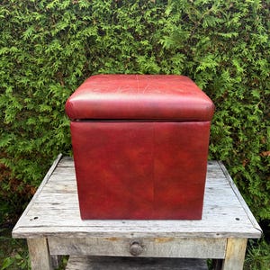 May include: A square, deep red storage ottoman with a smooth, leather-like finish. The top lid features a subtle, embossed starburst or compass-like geometric pattern centered on the surface. The sides are solid, flat, and uniform in color.