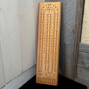 Könnte beinhalten: Ein hölzernes Cribbage-Spielbrett mit U-förmiger Bahn und mehreren Lochreihen für Stifte. Das Brett hat aufgedruckte Markierungen zum Zählen und den Text "INTERNATIONAL PLAYING CARD COMPANY LIMITED". Hergestellt in Taiwan.