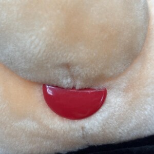 May include: Close-up of a plush toy face with a red, curved tongue. The toy's face is a light beige color, with soft, fluffy fur. The bottom portion of the image shows a black fabric.