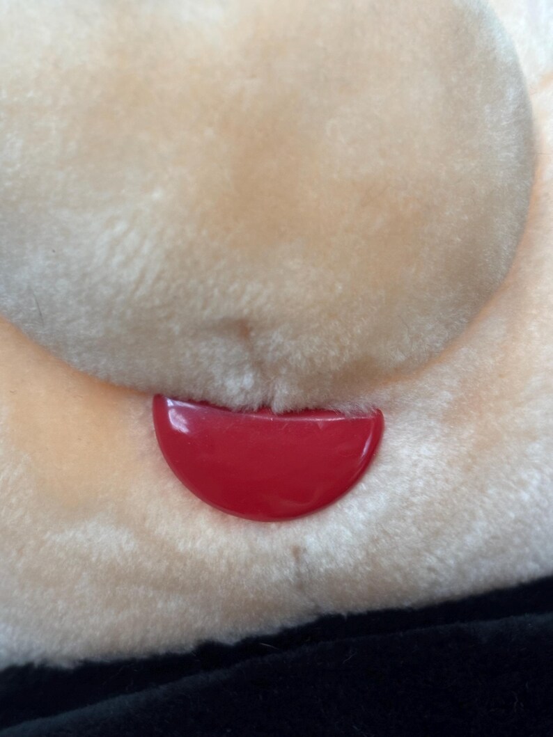 May include: Close-up of a plush toy face with a red, curved tongue. The toy's face is a light beige color, with soft, fluffy fur. The bottom portion of the image shows a black fabric.