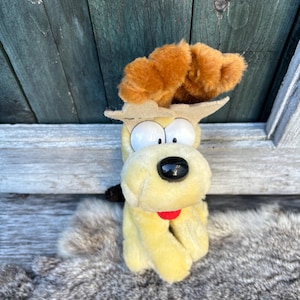 May include: A yellow plush dog toy with brown fur on its head. The dog has large black eyes, a black nose, and a red tongue. The toy is sitting on a grey furry surface.