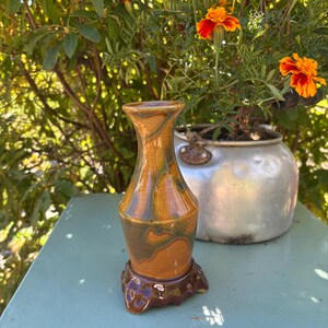 May include: A handmade ceramic vase with a brown and green marbled design. The vase is set on a dark brown, textured base. The background features orange flowers and foliage.