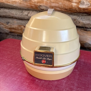 May include: Vintage Hoover portable hair dryer in cream and beige. The dome-shaped dryer has a handle and a gold-toned trim. The Hoover logo and product information are displayed on a rectangular plaque.