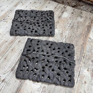 May include: Two rectangular, dark gray cast iron grates with a textured surface and numerous holes. Each grate has a thin, curved metal handle. The grates are displayed on a weathered wooden surface.
