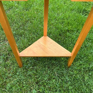 May include: A light brown wooden corner table featuring a triangular lower shelf and a single drawer with a small round wooden knob. The table is constructed with three straight legs that extend from the top frame to the base shelf.