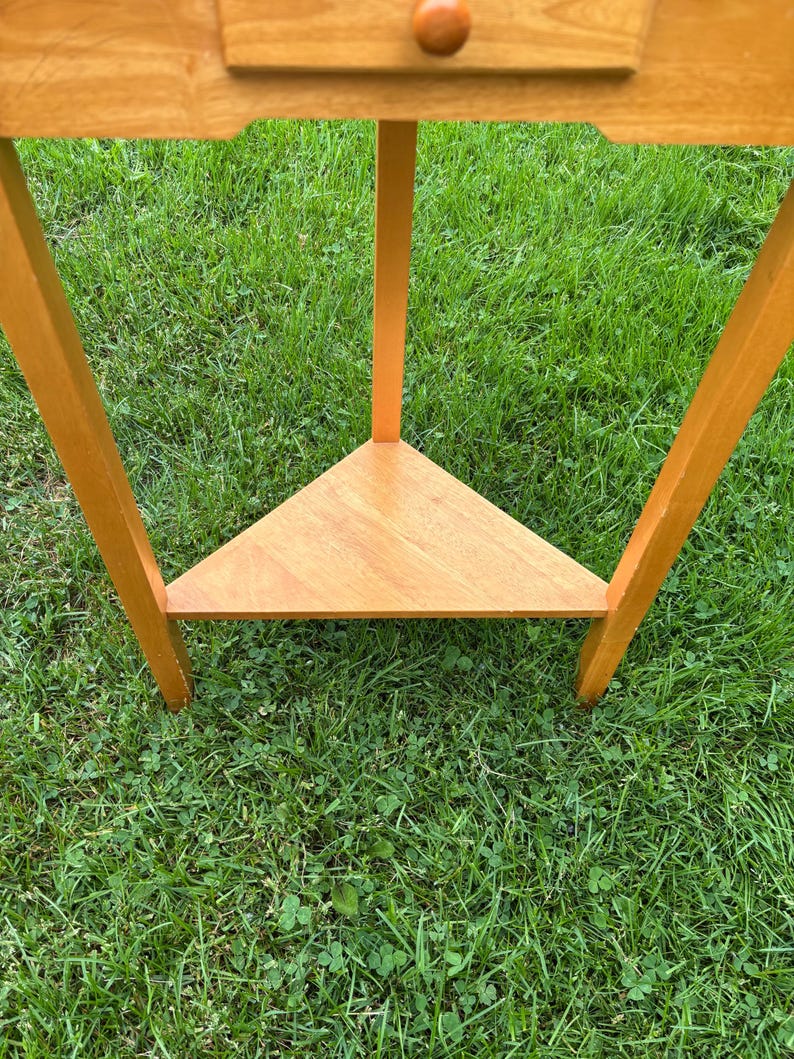 May include: A light brown wooden corner table featuring a triangular lower shelf and a single drawer with a small round wooden knob. The table is constructed with three straight legs that extend from the top frame to the base shelf.