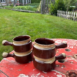 以下が含まれることがあります： 取っ手付きの、クリーム色の縁とリブ模様が特徴の、積み重ねられた2つの茶色のセラミック製スープボウル。 ボウルは、緑の芝生と温室を背景に、風化した赤い表面に置かれています。