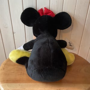 May include: A plush Minnie Mouse doll, viewed from the back, with black fur, large round ears, and a red bow. The doll has yellow shoes and white gloves. The doll is sitting on a wooden surface.