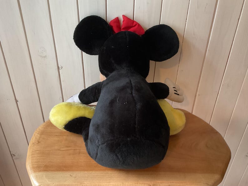 May include: A plush Minnie Mouse doll, viewed from the back, with black fur, large round ears, and a red bow. The doll has yellow shoes and white gloves. The doll is sitting on a wooden surface.