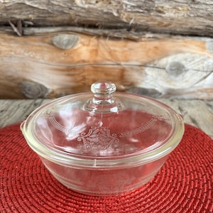 May include: A clear glass casserole dish with a matching lid. The dish has a round shape with a decorative etched design. The lid has a small knob handle. The dish sits on a red woven mat against a wooden background.