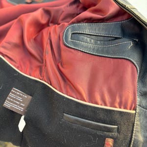 May include: Close-up of the inside of a black leather jacket. The lining is a dark red color with a white trim. The jacket has a black leather pocket on the inside. The label reads "Made in India.  Fit in size. Designed in Canada.  Conception Canadienne. Style No./Catalogue # 800057. Professional Leather Cleaning/Nettoyage professionnel du cuir/lavaggio professionale della pelle."