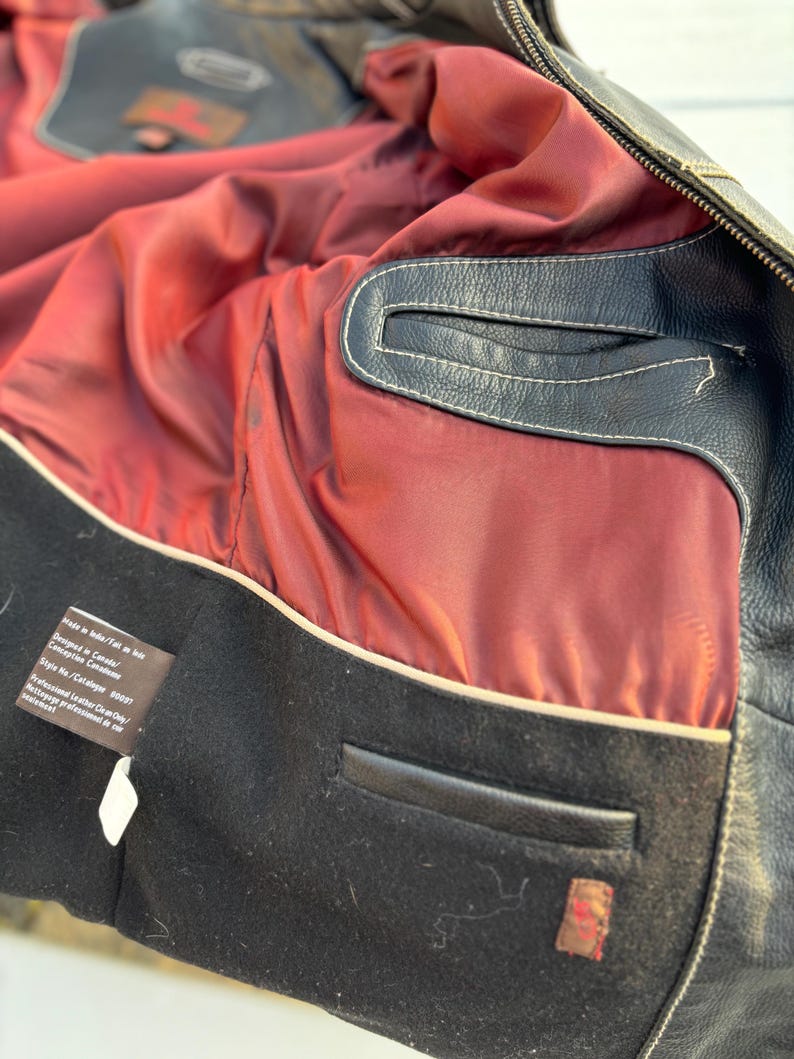 May include: Close-up of the inside of a black leather jacket. The lining is a dark red color with a white trim. The jacket has a black leather pocket on the inside. The label reads "Made in India.  Fit in size. Designed in Canada.  Conception Canadienne. Style No./Catalogue # 800057. Professional Leather Cleaning/Nettoyage professionnel du cuir/lavaggio professionale della pelle."