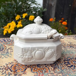 May include: A white ceramic sugar bowl with an octagonal shape, featuring embossed fruit motifs including grapes and peaches on the sides and lid. The bowl includes a matching lid with a central finial and a small ceramic spoon resting inside.