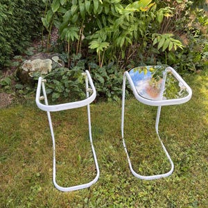 Puede incluir: Dos mesas auxiliares de metal blanco con tapas de cristal se colocan sobre un césped. Las mesas tienen un diseño de patas curvas y se colocan contra un fondo de exuberante follaje verde. Las tapas de cristal reflejan el cielo.