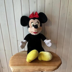 May include: A plush Minnie Mouse doll, with a black body, large black ears, and a red bow. The doll has white gloves and yellow shoes. The doll is sitting on a wooden table.