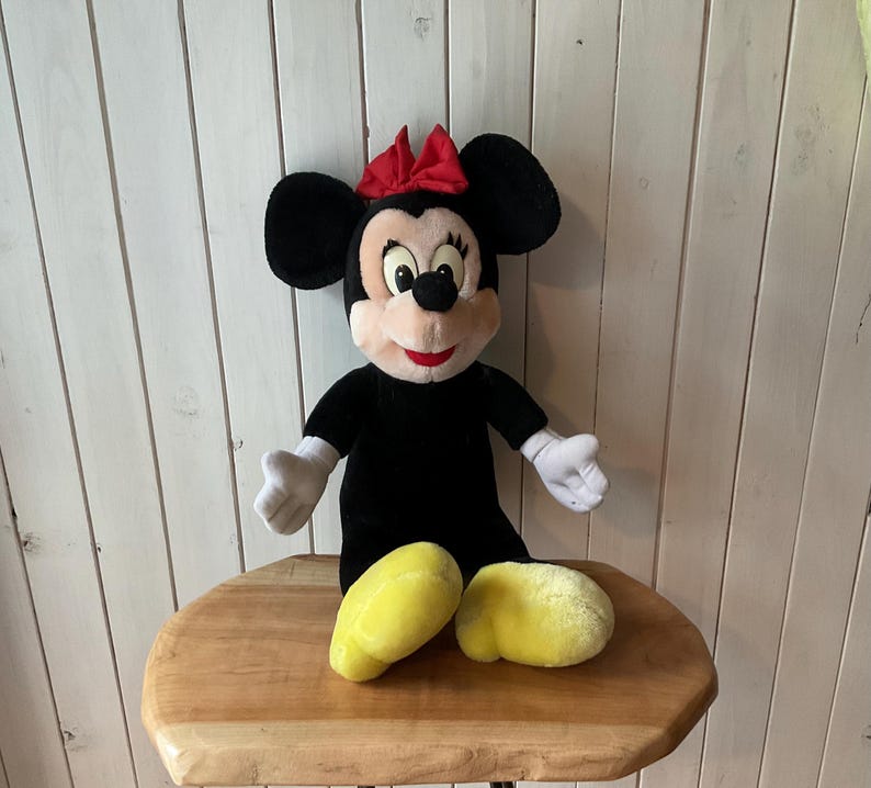May include: A plush Minnie Mouse doll, with a black body, large black ears, and a red bow. The doll has white gloves and yellow shoes. The doll is sitting on a wooden table.