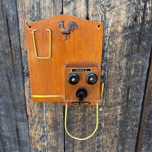 May include: A vintage wooden telephone index with a brass rooster emblem. The index features a cradle for a handset, a pen holder, and the text "SUPPORT TELEPHONE". The wooden frame has a warm, reddish-brown hue.