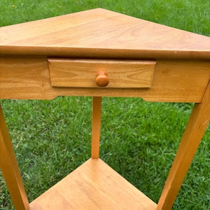 May include: A light brown wooden corner table featuring a triangular top, a single drawer with a small round knob, and a lower triangular shelf. The table is supported by three straight legs and has a smooth, natural wood grain finish throughout.
