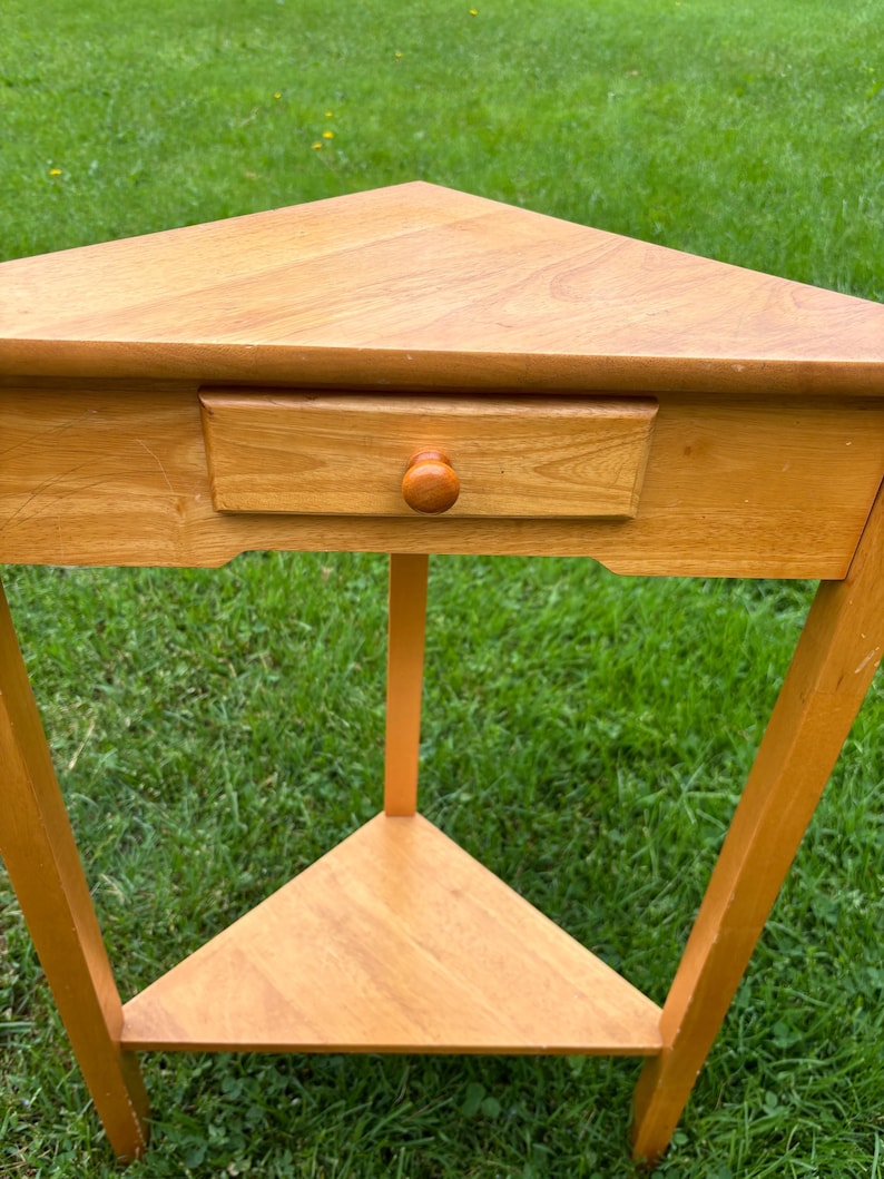 May include: A light brown wooden corner table featuring a triangular top, a single drawer with a small round knob, and a lower triangular shelf. The table is supported by three straight legs and has a smooth, natural wood grain finish throughout.