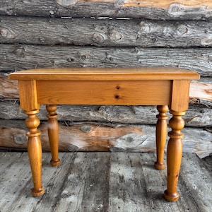 May include: A small, rectangular wooden table with a warm, honey-toned finish. The table features a single drawer and four turned legs. The table's dimensions are approximately 76 cm long, 46 cm wide, and 71 cm high.